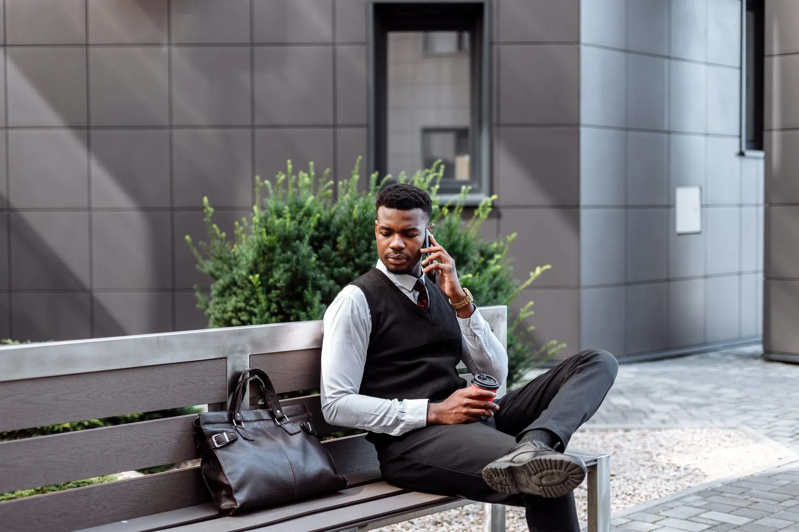 Stylish businessman talking on phone while holding coffee on a bench outdoors.